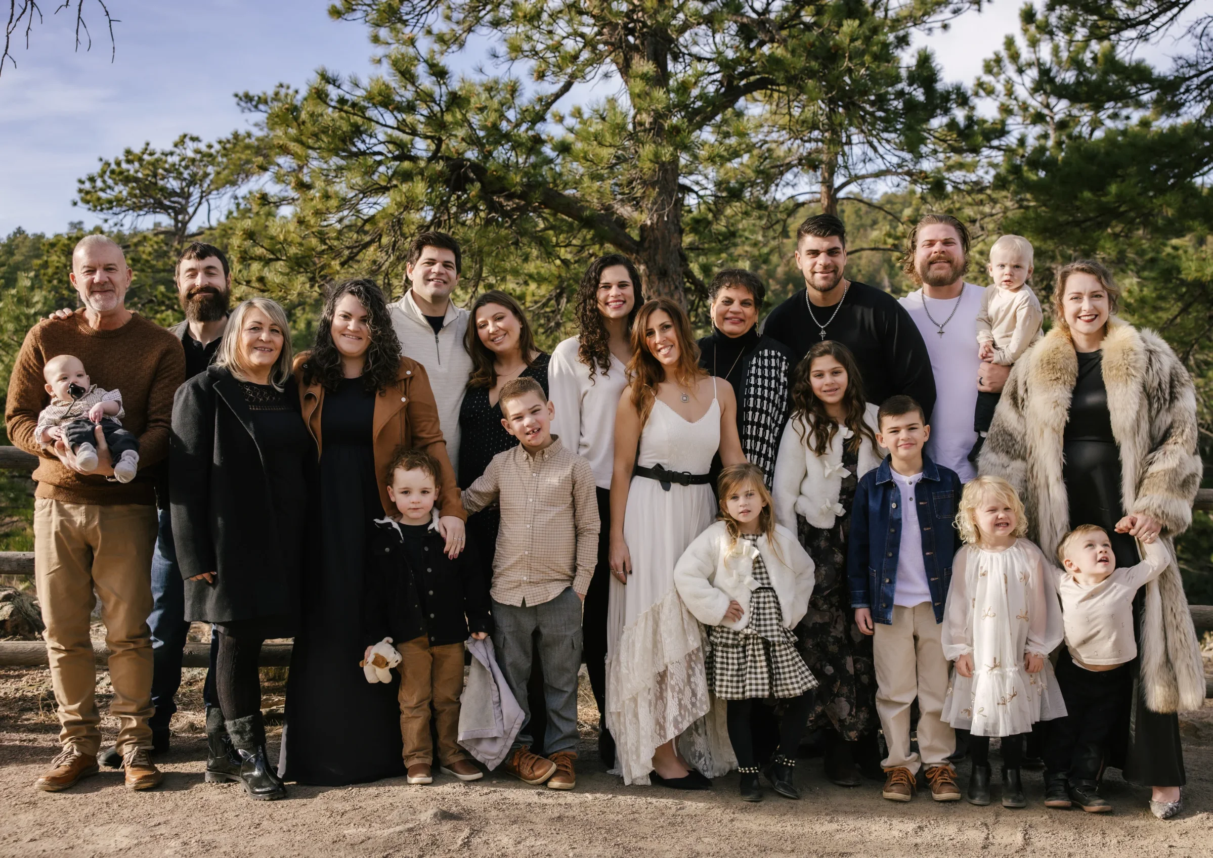 Large family group outdoors posing together in front of pine trees on a sunny day.
