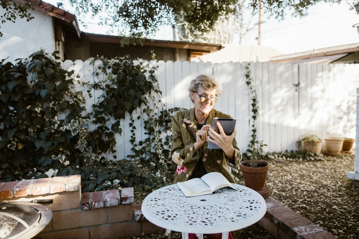 Smiling elderly woman using a tablet while sitting at a white metal outdoor table with an open book in a garden.