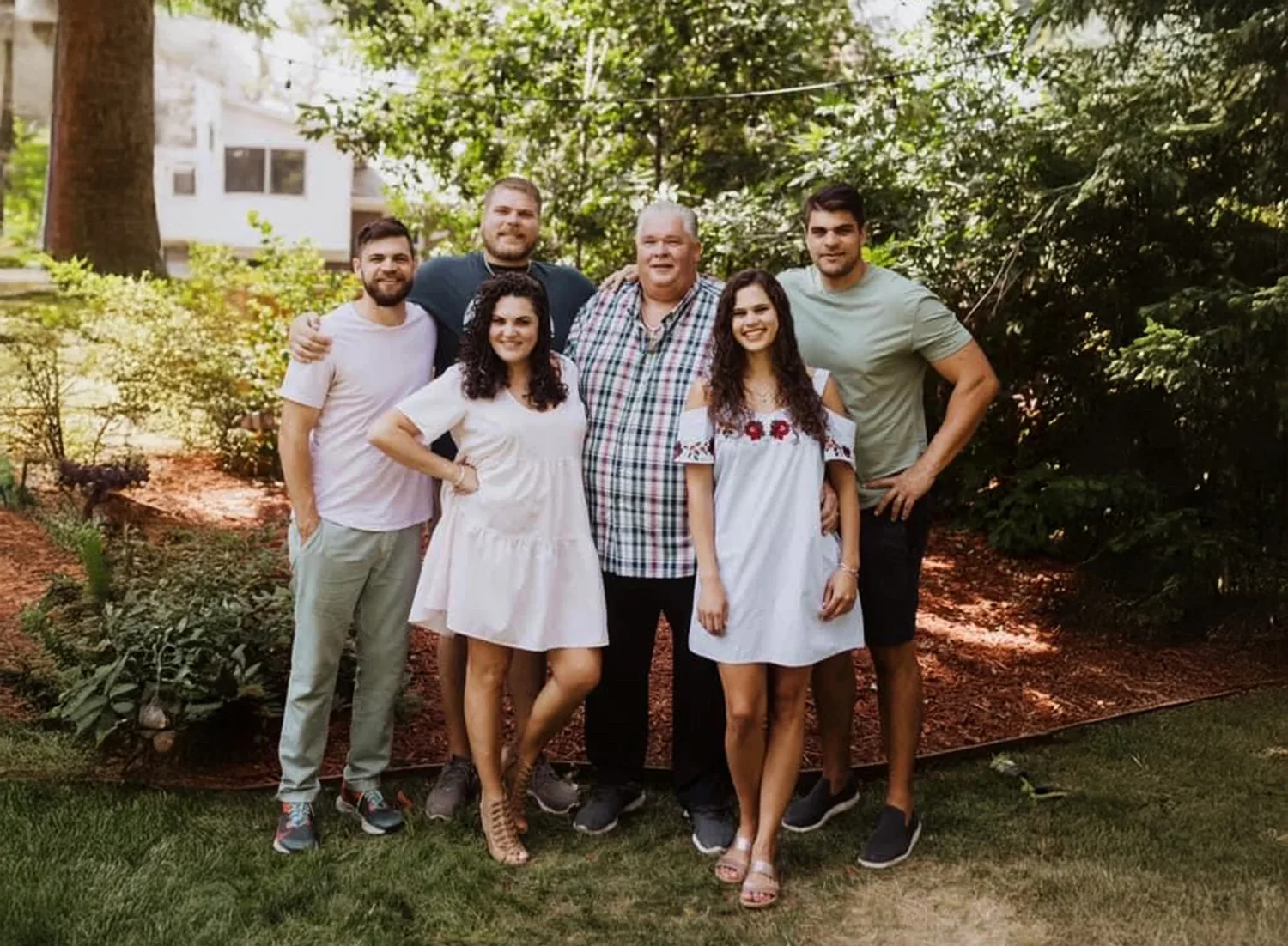 Group of six people posing outdoors on grass with trees and bushes in the background, including two women in white dresses and four men in casual clothing.