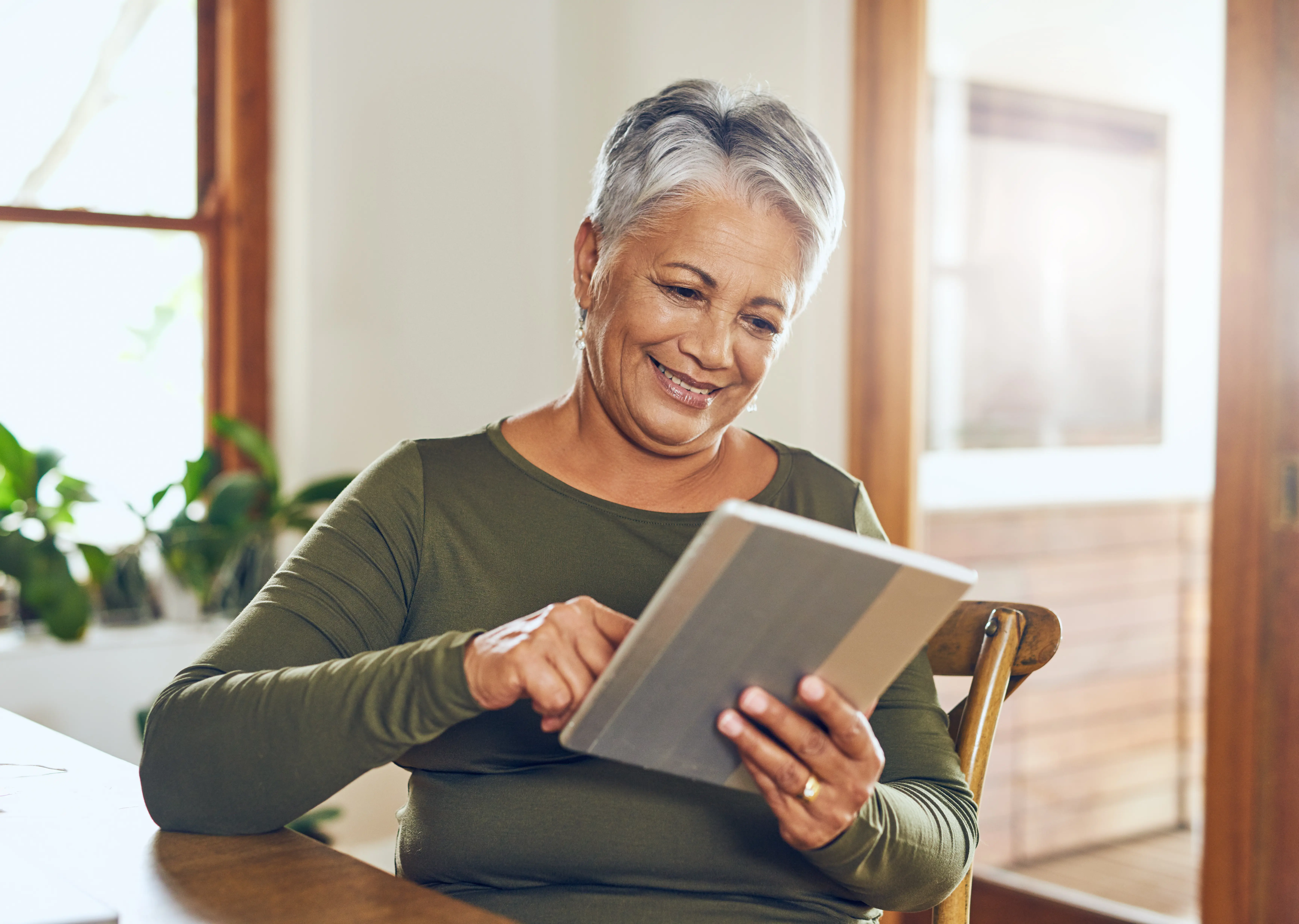 Smiling older woman with short gray hair using a tablet at a wooden table in a bright room.