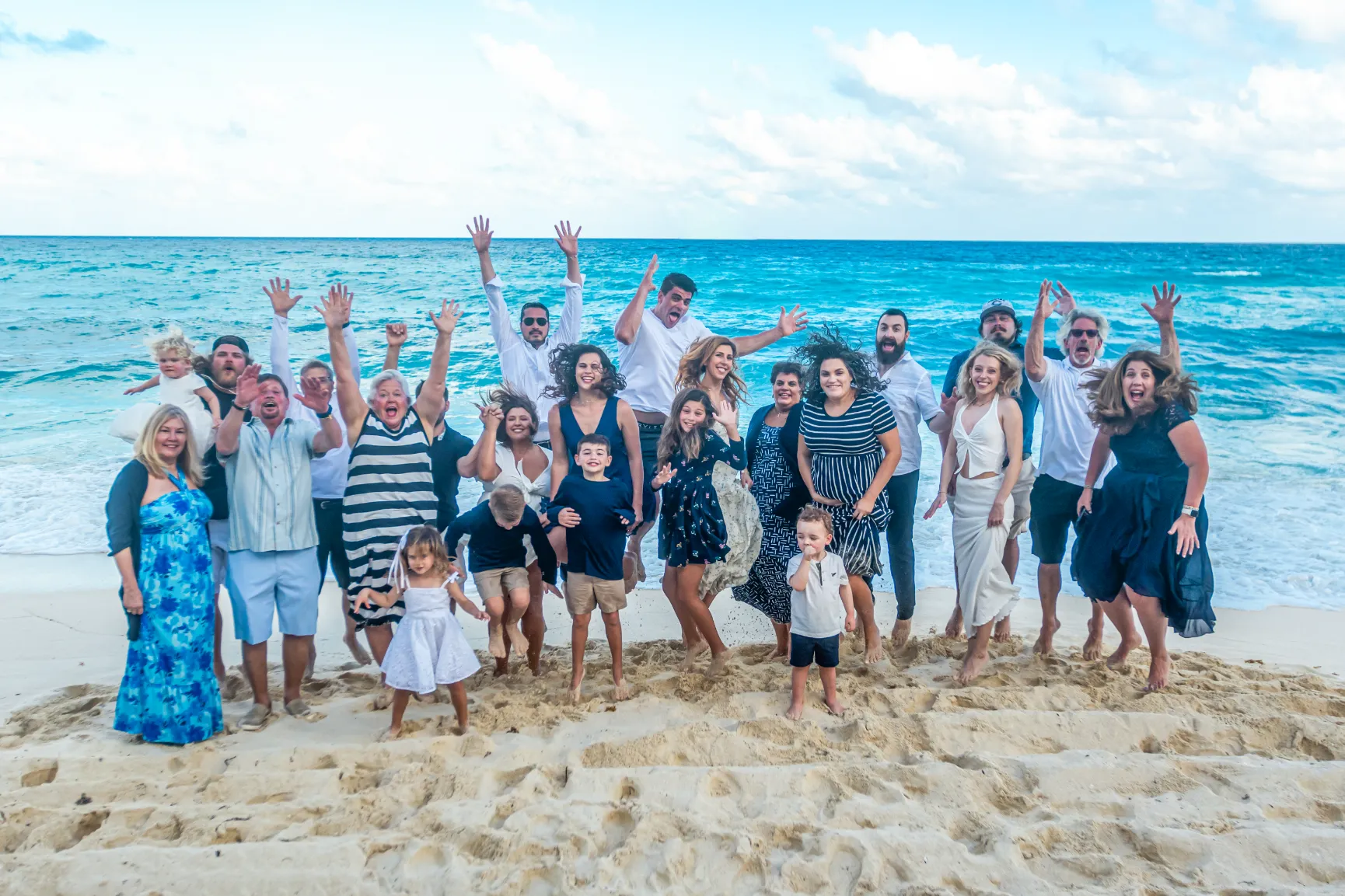 Large family jumping and smiling together on a sandy beach with blue ocean and cloudy sky in the background.