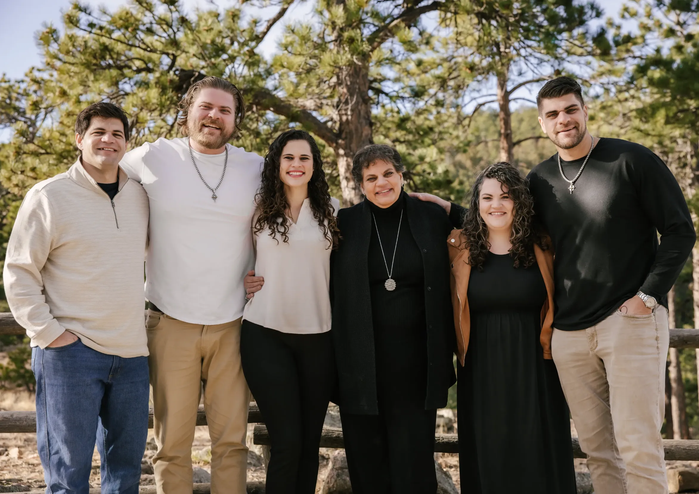 Group of six adults standing outdoors with arms around each other in front of trees, smiling at the camera.