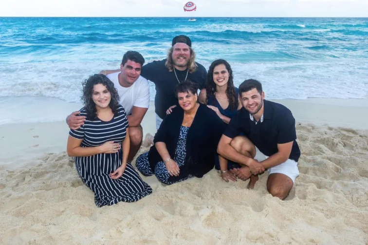 A woman and five adults sitting closely together on a sandy beach with ocean waves in the background, smiling at the camera.
