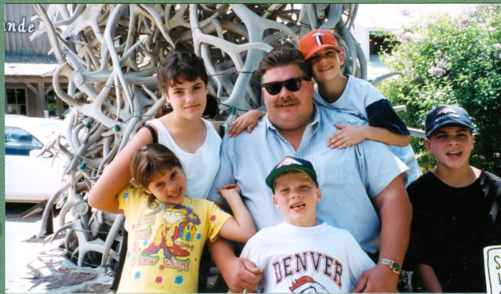 A man wearing sunglasses and four kids standing closely together outdoors in front of a large display of intertwined antlers.